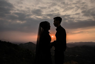 Couple standing on mountain against sky during sunset