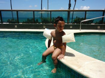 Shirtless boy looking away while sitting at poolside during sunny day