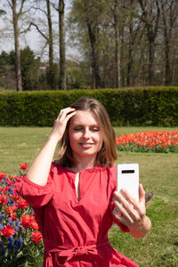 A young beautiful girl in a red dress in a spring park plays and takes a selfie