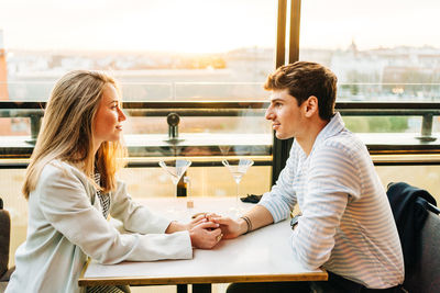 Side view of loving couple in trendy wear sitting at table in rooftop restaurant while holding hands and looking at each other