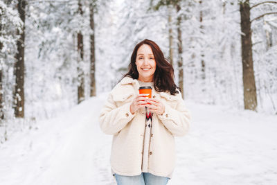 Smiling woman in winter clothes with paper cup of coffee in hands in snowy winter forest