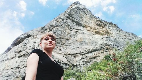 Low angle portrait of young woman standing on cliff against sky