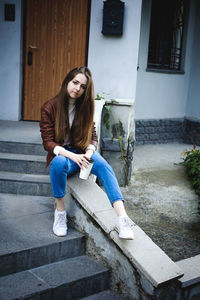 Portrait of young woman holding coffee cup while sitting against house