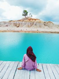 Rear view of woman sitting on rock by lake against sky