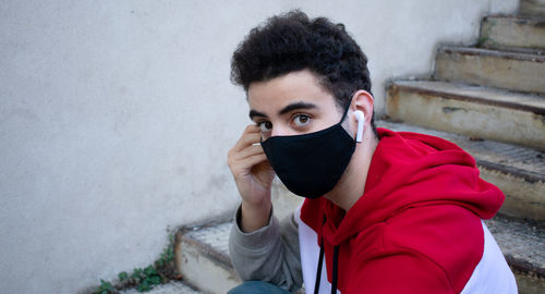 Close-up of a young caucasian man putting an earbud, while sitting on some stairs, with face mask