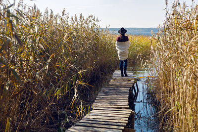 Woman in a black hat and sweater stands on the old pier to the bushes by the lake