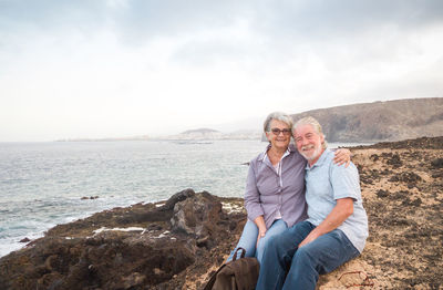 Portrait of smiling senior couple sitting on rocky coastline