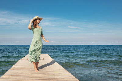 A lovely slender caucasian woman in a green dress and a straw hat is walking along the pier 