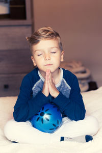 Smiling boy with eyes closed praying while sitting with ball on bed at home