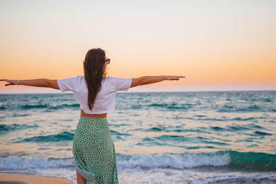 Rear view of woman standing at beach against clear sky during sunset