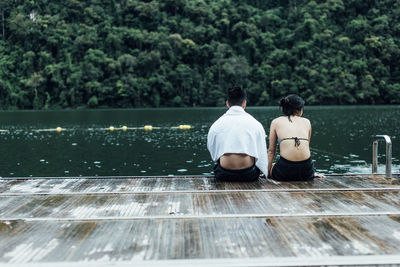 Rear view of couple sitting by lake against trees