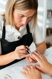 High angle of crop concentrated female manicurist in apron applying polish on nails of unrecognizable client sitting at table in modern beauty salon