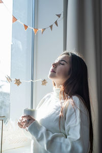 Woman in warm white winter sweater sitting on the window at home at christmas eve holding cup