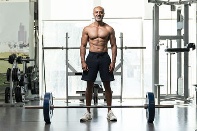 Full length of shirtless man with barbell standing in gym