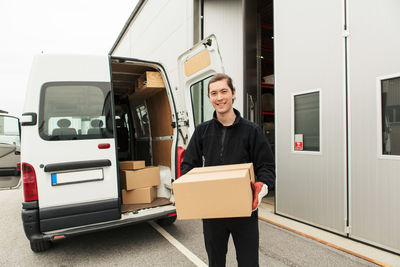 Portrait of smiling manual worker carrying cardboard box while standing against delivery van