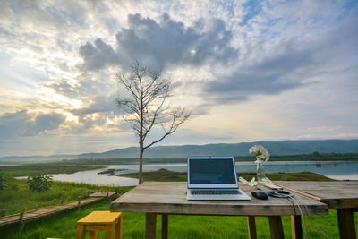 Laptop on table at lakeshore against cloudy sky during sunset