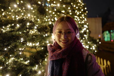 Portrait of young woman standing by christmas tree at night