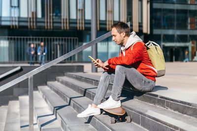Hipster with wooden longboard sitting on the steps and chatting in mobile phone. leisure activity