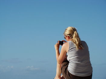 Rear view of woman photographing with mobile phone against blue sky