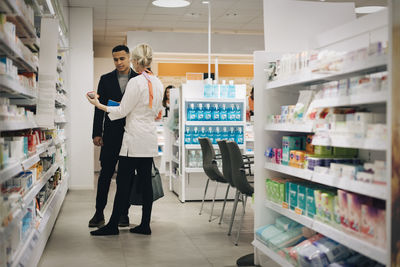 Full length of mature female pharmacist showing medicine to male customer standing by rack at store