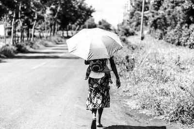 Rear view of woman with umbrella carrying baby while walking on street during sunny day
