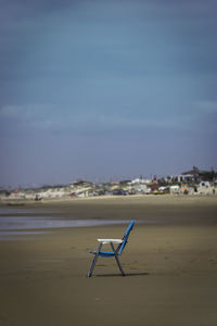 Empty chair on sand at beach against blue sky