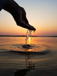 Cropped hand in sea against sky during sunset