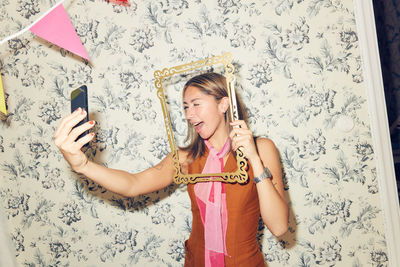 Cheerful young woman taking selfie through picture frame while winking at smart phone against wallpaper during party in