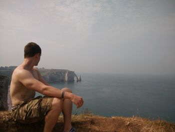 Shirtless man looking at sea while sitting on rock against sky