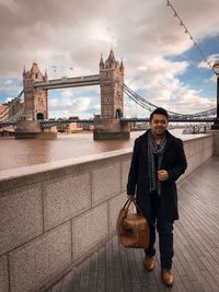 Full length portrait of young man standing by thames river with tower bridge in background