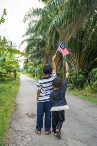 Rear view siblings holding malaysian flag while standing on road amidst plants