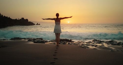Rear view of woman standing at beach against sky during sunset