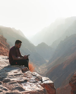 Young man sitting on mountain against sky