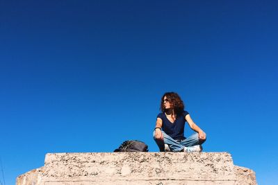 Low angle view of woman sitting on rock against blue sky