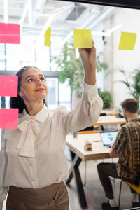 Happy female worker taking notes on stickers attached on glass wall of room with colleagues while working in modern office