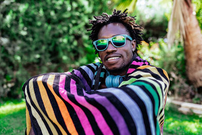 Smiling african american male in sunglasses wrapped in colorful towel sitting on meadow in summer park and looking at camera