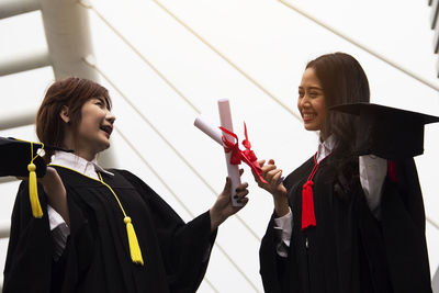 Cheerful young women holding degree certificates standing against sky