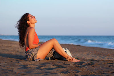 Young woman sitting at beach against sky during sunset