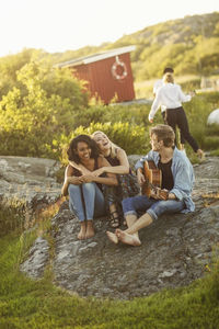 Man playing guitar while friends enjoying on rock formation during summer