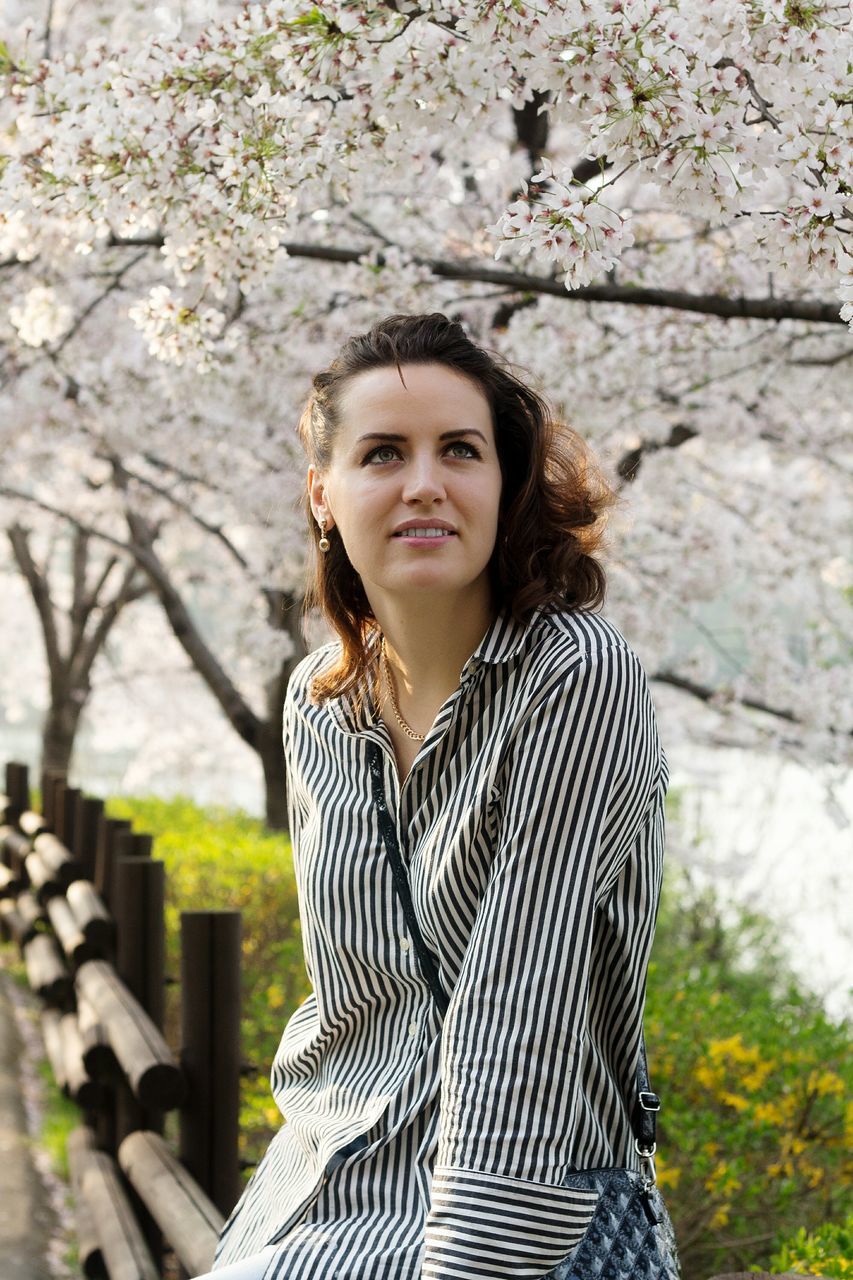 tree, young adult, real people, young women, looking at camera, one person, leisure activity, front view, casual clothing, portrait, focus on foreground, day, outdoors, lifestyles, standing, branch, beautiful woman, flower, growth, nature, close-up