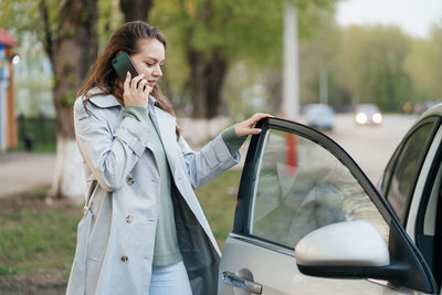 Beautiful girl with long hair in a grey trench coat using smartphone call gets into the car