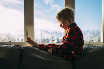 Side view of boy looking at decor while sitting on sofa by window at home