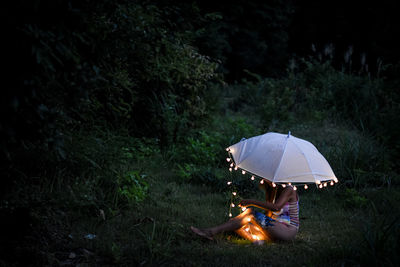 Woman sitting on field by trees in forest with an umbrella and fairy lights 