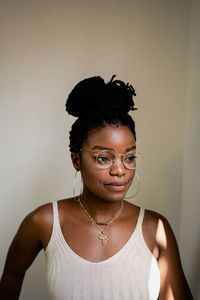 African american female in white top and round transparent eyeglasses with dark hair tied up looking away while standing in room near grey wall