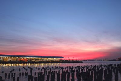 Wooden posts in sea by illuminated pier against sky at dusk