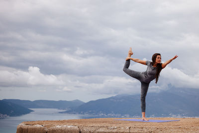 Full length of young woman exercising on cliff against sky