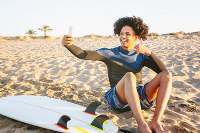 African american man sitting on the sand with surfboard takes a photo with a smartphone