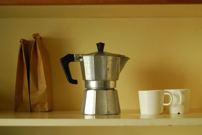 Close-up of coffee maker with cups and paper bags on shelf