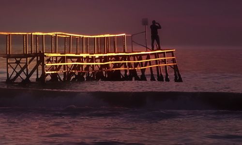 Man working on bridge over sea against sky during sunset