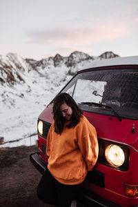 Young happy tourist in eyeglasses standing near vintage automobile between deserted ground in snow near mountains